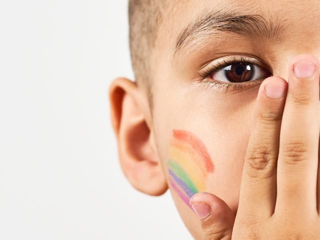 small boy of unknown ethnicity covering mouth with hand and a rainbow painted on his cheek